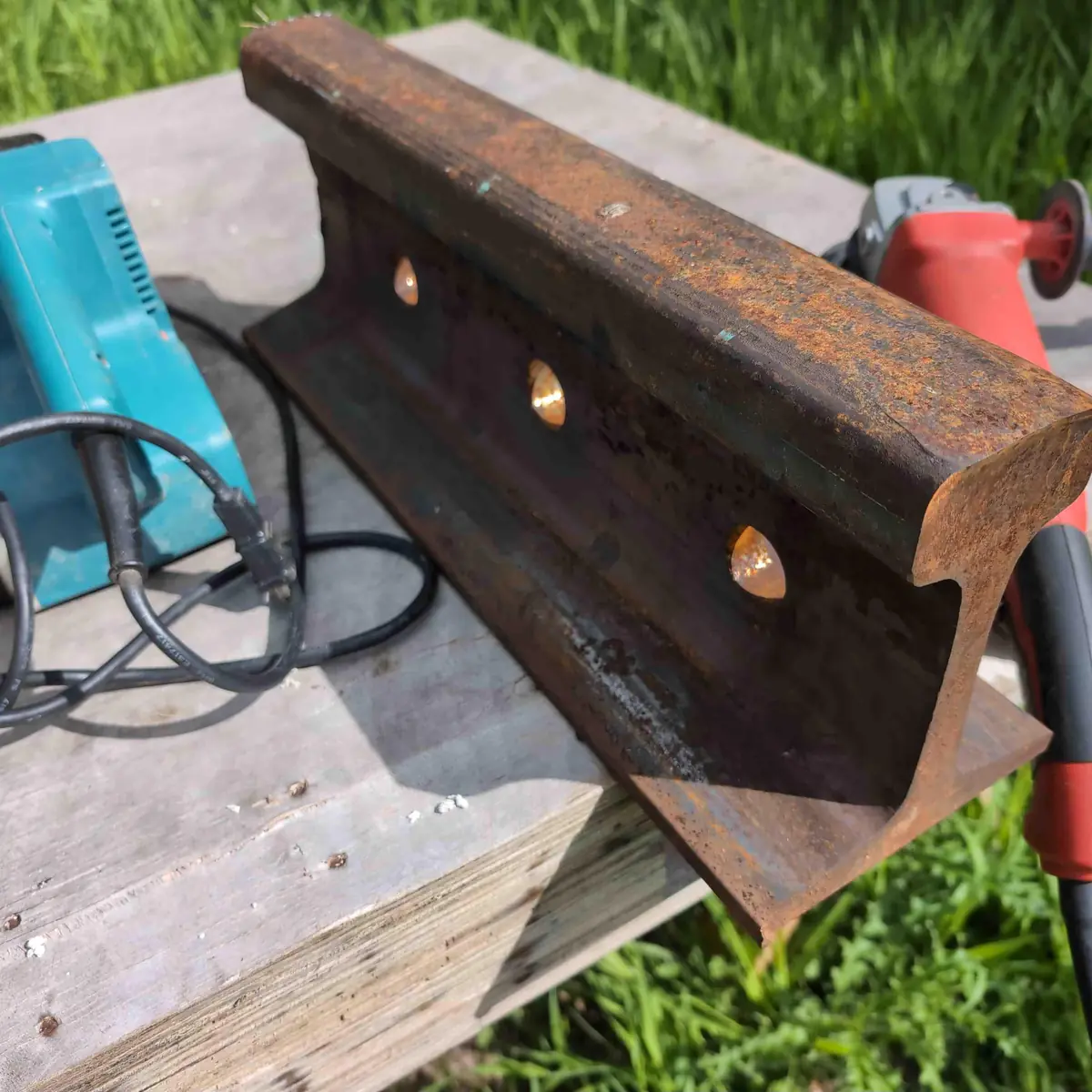 A rusty chunk of rail track with an angle grinder and belt sander next to it.